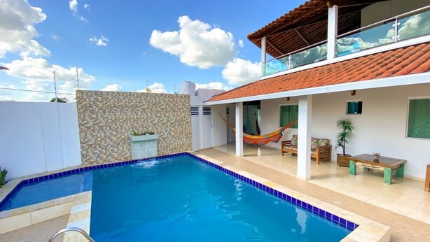 Una piscina con acqua cristallina blu, circondata da un muro bianco con motivo in pietra, un pontile in legno e un'area relax coperta con un'amaca.