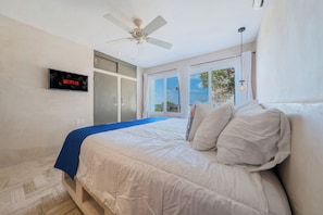 Ein Schlafzimmer mit einem Bett, einem Deckenventilator, einem Fernseher, der das Netflix-Logo zeigt, und einem Fenster mit Vorhängen.