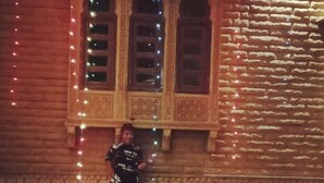 A person standing in front of a building with a stone facade and wooden window frames, decorated with string lights. - Blessing Hotel (Jaisalmer)