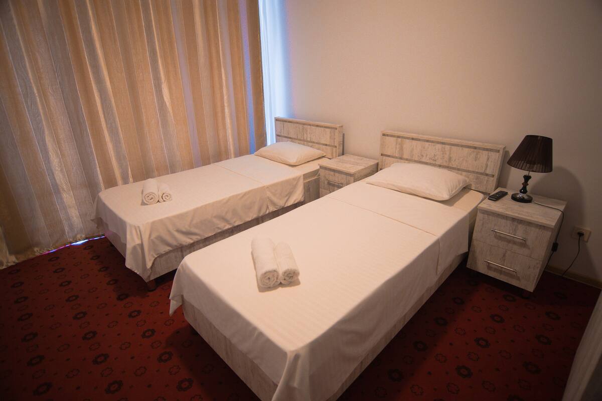A hotel room with two single beds, a nightstand with a lamp, and a window with curtains.