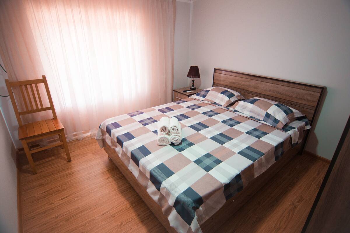 A neatly made bed with a checkered bedspread, a wooden nightstand with a lamp, and a wooden chair.