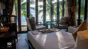 A hotel room with a large bed, a small table, two chairs, and a view of the beach and palm trees through large windows. - Victoria Cliff Nyaung Oo Phee Island (Kawthaung)