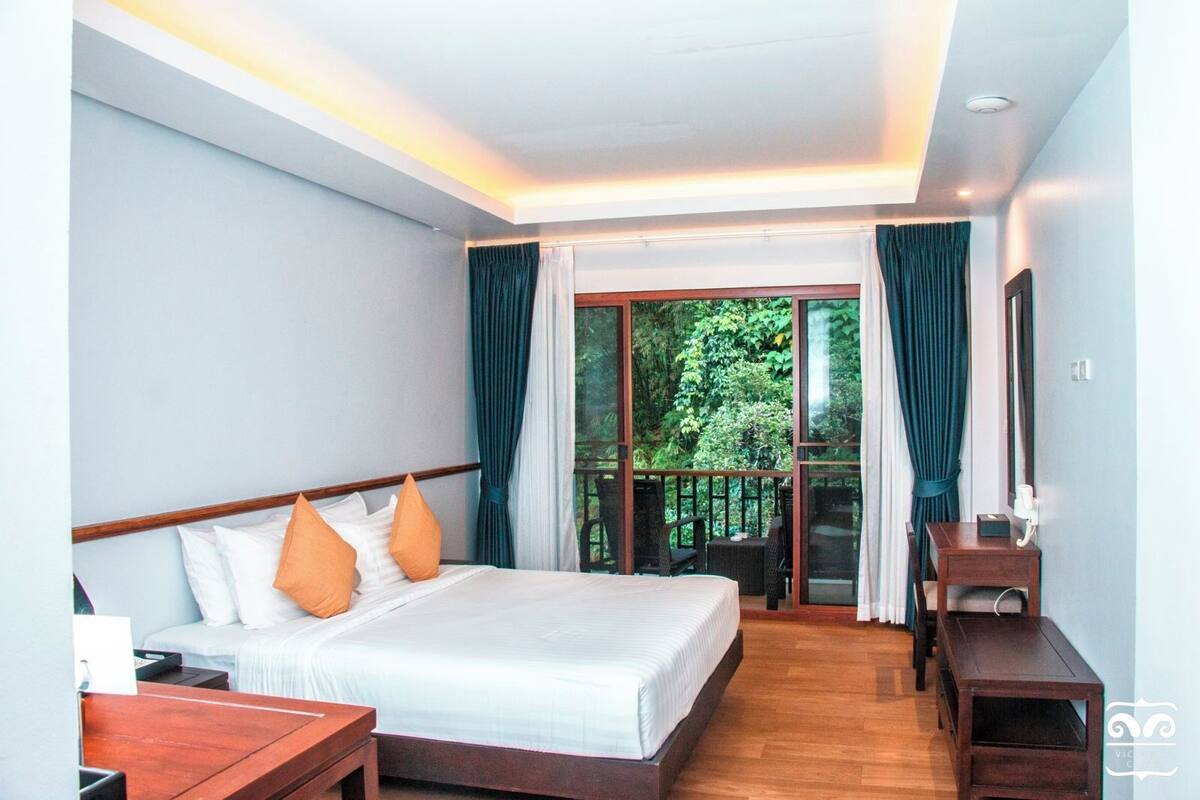 Ein Hotelzimmer mit Bett, Nachttischen aus Holz, einem Schreibtisch und Blick ins Grüne durch große Fenster.