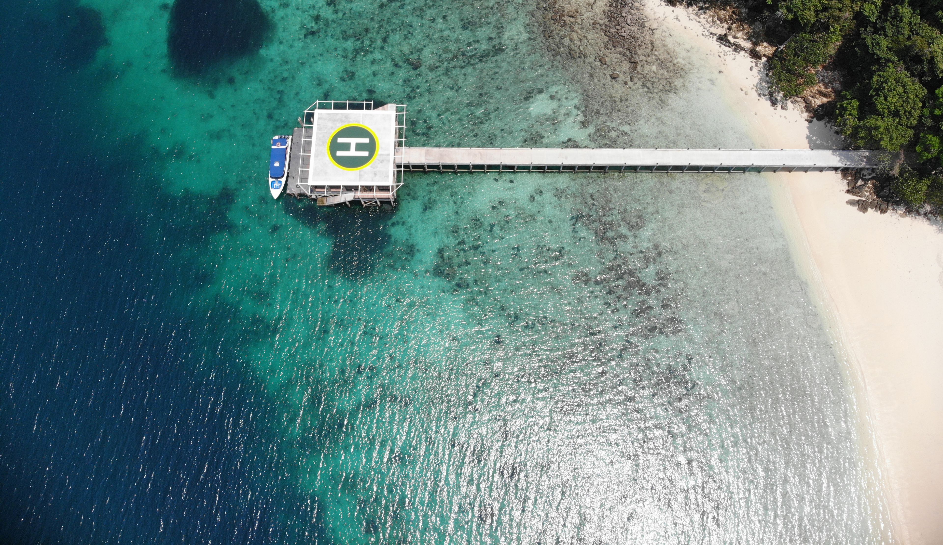 Una vista aerea di una spiaggia con un molo che si protende sull'oceano, una barca ormeggiata al molo e un elisuperficie sul molo.