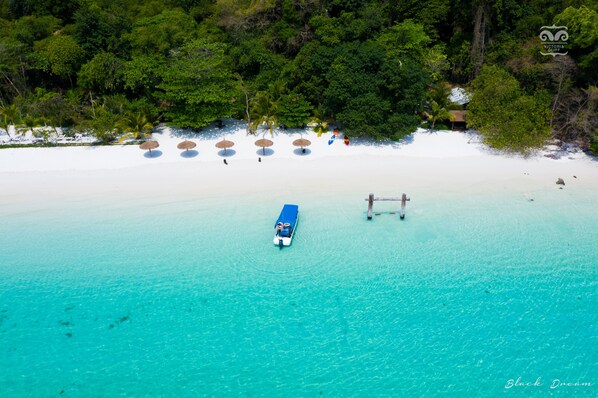 Vue aérienne d’une plage aux eaux turquoise et limpides, avec un bateau et plusieurs parasols de plage.