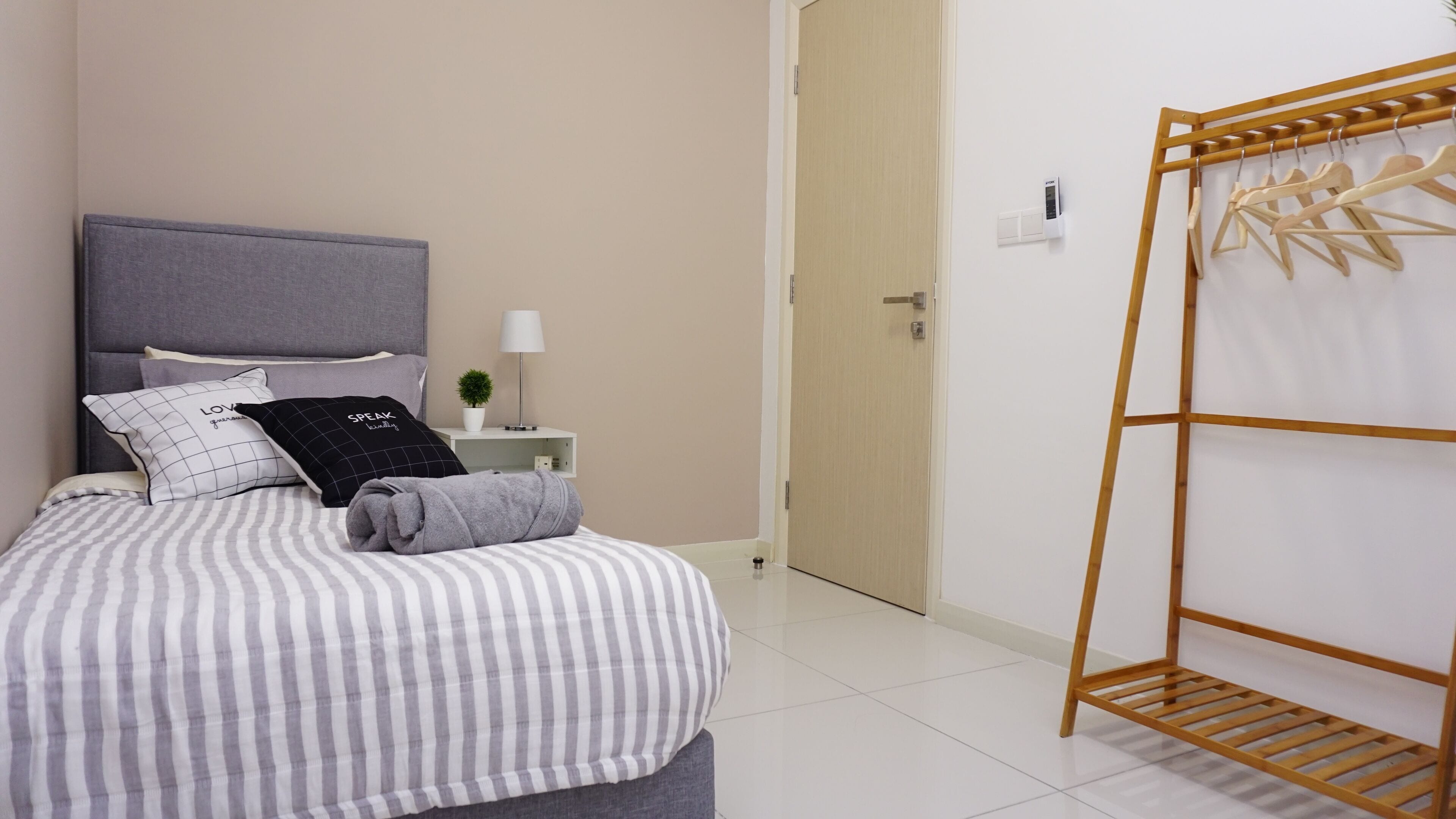 A neatly made bed with striped bedding, a grey headboard, and two pillows with text. A small bedside table with a lamp and a potted plant is beside the bed. A wooden drying rack with clothes hangs on the wall.