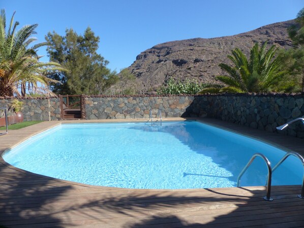 A rectangular swimming pool with a stone wall and wooden deck, surrounded by trees and a mountain in the background.