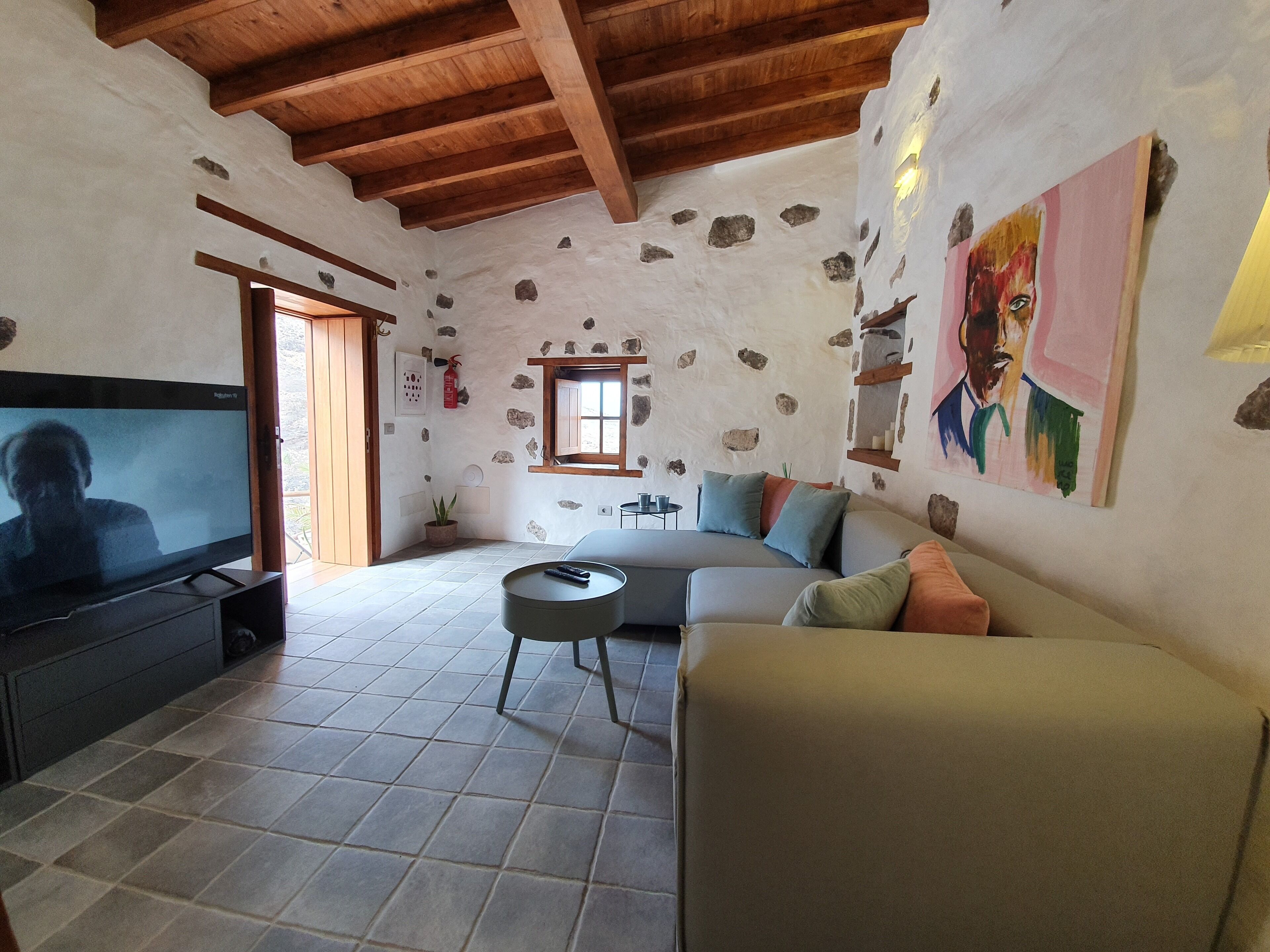Una sala de estar acogedora con pared de piedra, vigas de madera en el techo, un televisor de pantalla plana, un sofá y una pequeña mesa redonda.