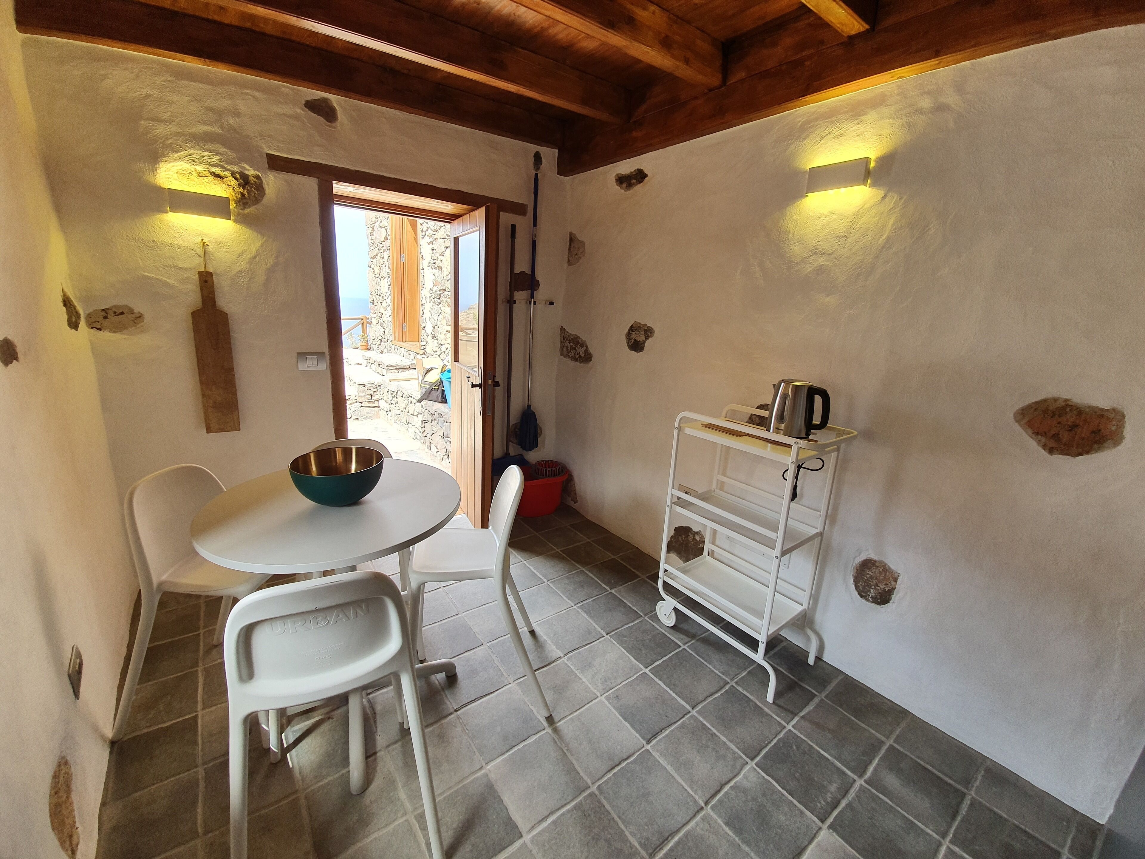 A small dining area with a table and chairs, a white wall with stone patches, a wooden ceiling, and a view through an open door.