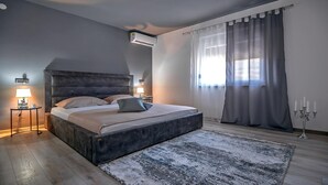 Une chambre moderne avec un grand lit, des lampes de chevet, un climatiseur au plafond et une vue sur l’extérieur à travers des rideaux légers.