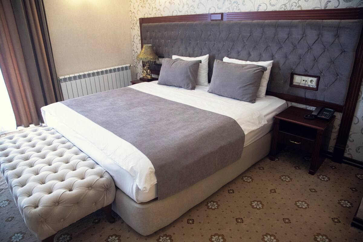 A neatly made bed with a grey bedspread and pillows, a wooden nightstand with a telephone, and a tufted bench beside the bed.