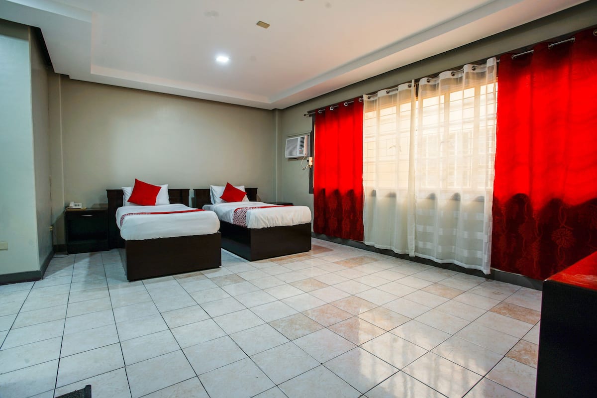 A hotel room with two beds, a flat-screen TV, and a window with red curtains.
