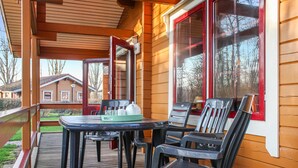 Eine Holzterrasse mit Tisch und Stühlen, ein überdachter Bereich mit einem Fenster und Blick auf ein Haus im Hintergrund.