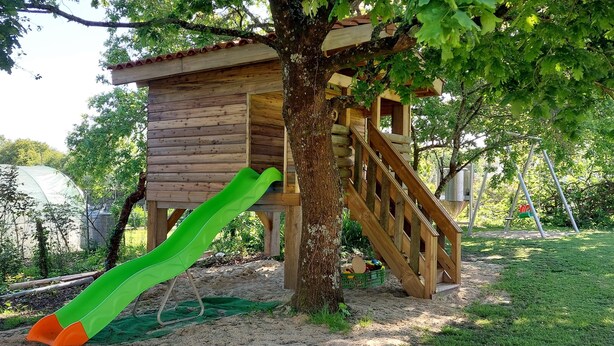 Una casita de juegos de madera con tobogán y escaleras, rodeada de árboles y césped.