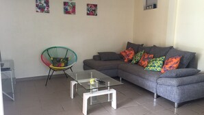 Ein modernes Wohnzimmer mit einem grauen Sofa, farbenfrohen Kissen, einem Glastisch und einem Korbstuhl.