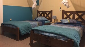 Ein Zimmer mit zwei Holzbetten, jeweils mit blauer Steppdecke und einem Holz-Kopfteil. An einer Seite befindet sich ein kleiner Nachttisch mit einer Lampe, an der anderen eine wandmontierte Steckdose.