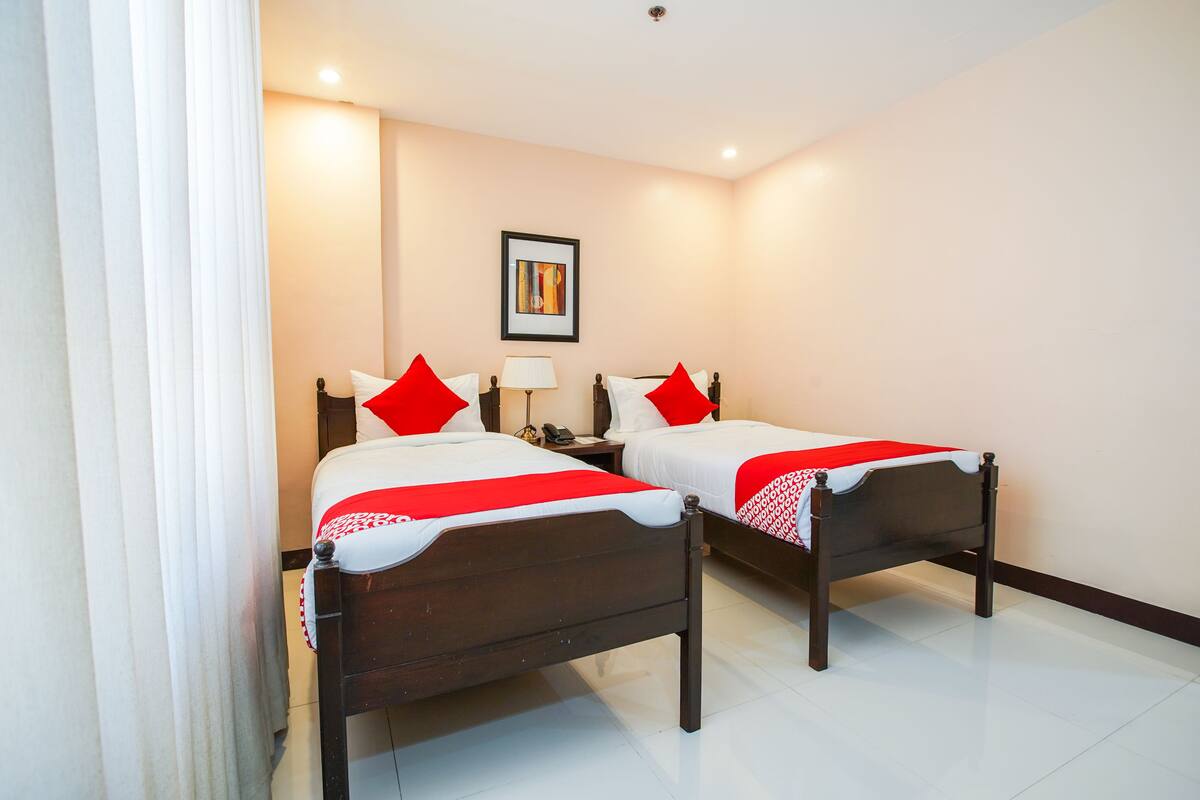 Une chambre d’hôtel avec deux lits en bois, du linge de lit blanc, des oreillers rouges, une table de chevet avec une lampe et un tableau encadré au mur.