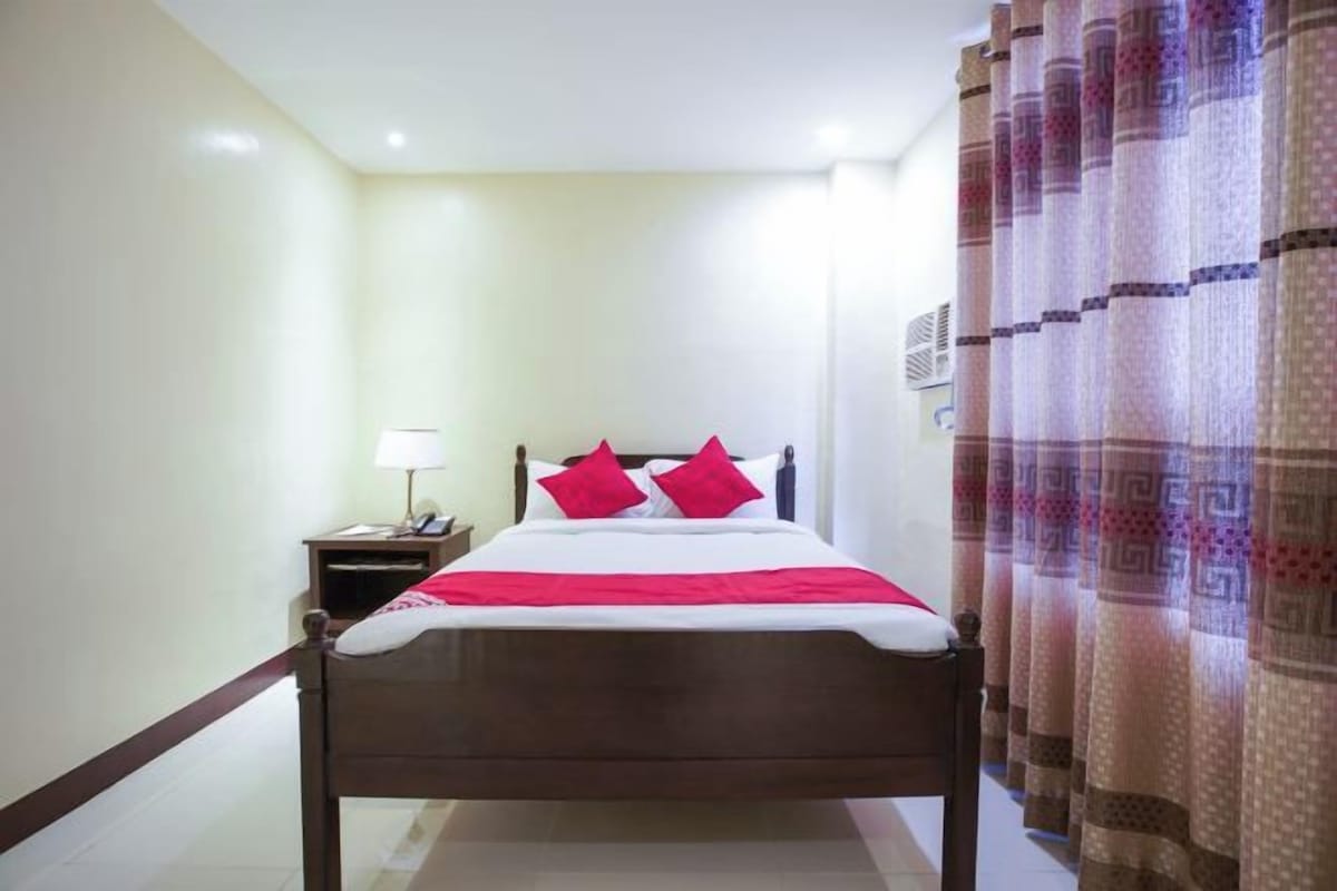 Habitación de hotel con cama, mesita de noche, lámpara y cortinas.