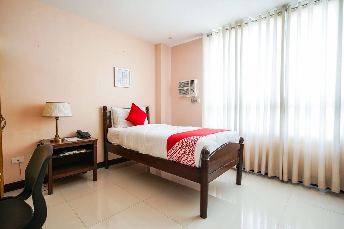 Une chambre d’hôtel équipée d’un lit, d’un bureau, d’une chaise, d’une lampe, d’une fenêtre avec des rideaux et d’un appareil de climatisation.