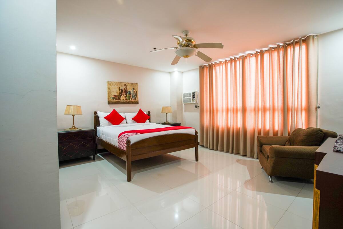 Une chambre d’hôtel avec un lit, un ventilateur de plafond, une fenêtre avec des rideaux, une table de chevet, une lampe et une chaise.