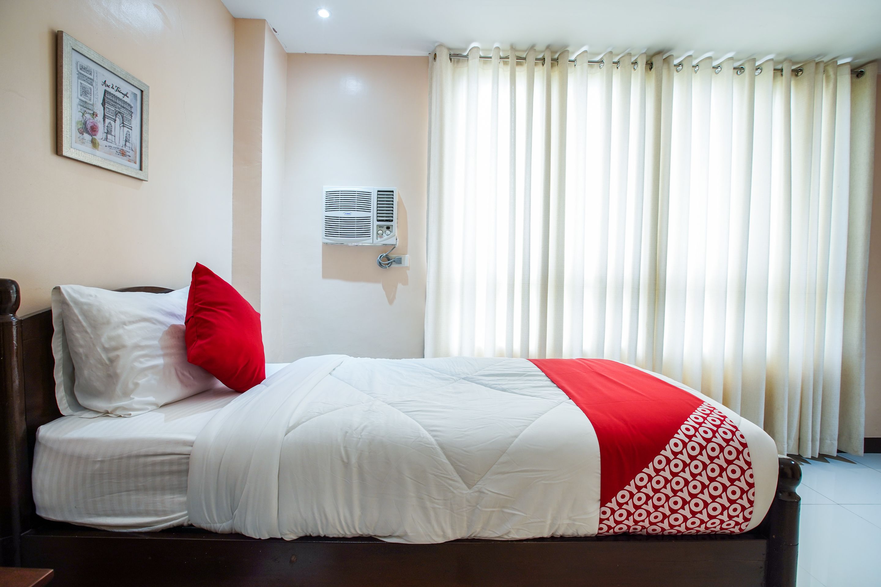 A neatly made bed with white and red bedding, a wall-mounted air conditioner, and a window with vertical blinds.