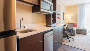 Una habitación de hotel moderna con kitchenette, que incluye un refrigerador de acero inoxidable, microondas y lavavajillas. Hay un escritorio con una silla y una lámpara, y una pequeña zona de estar con un sofá y reposapiés.