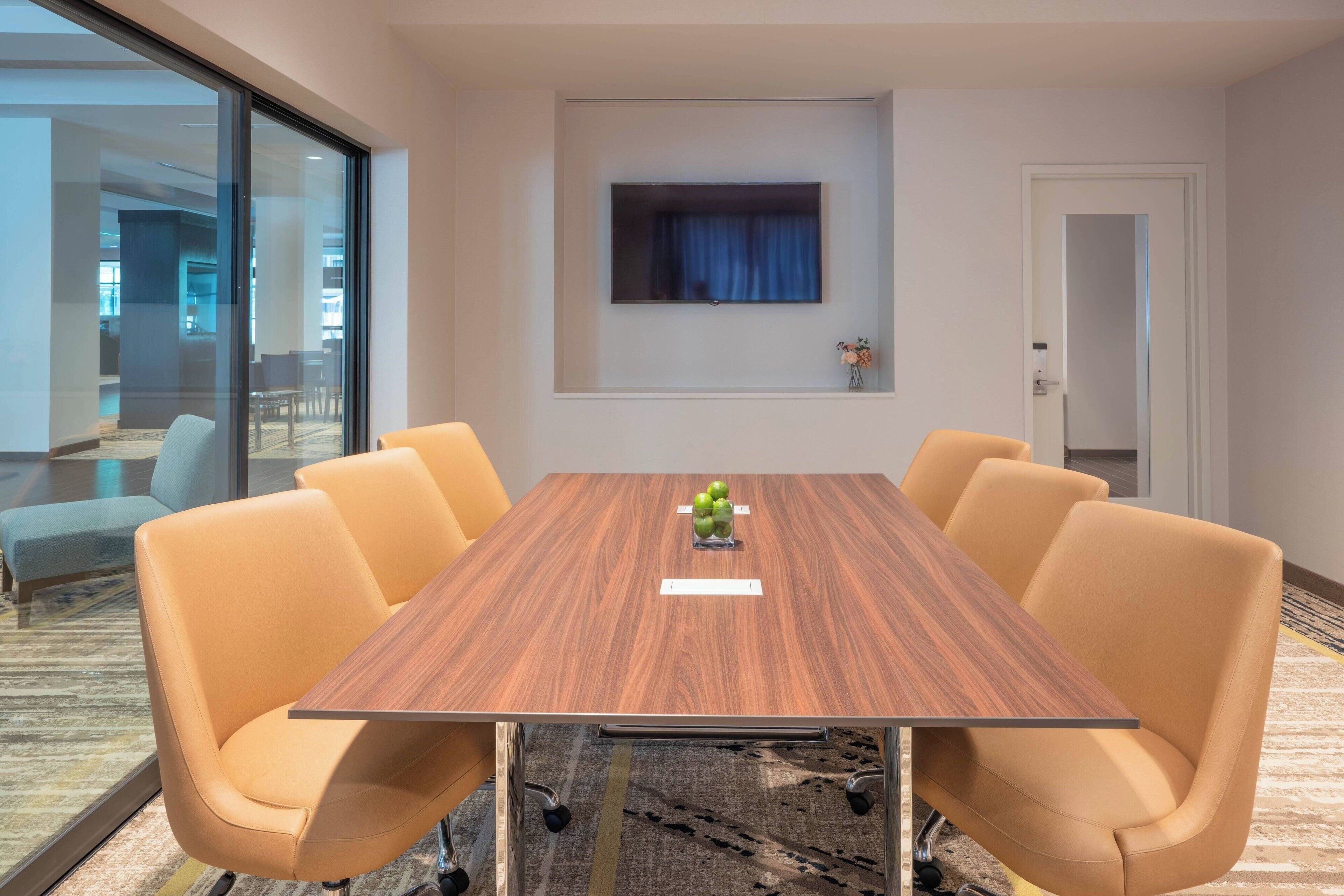 A conference room with a wooden table, four orange chairs, a flat-screen TV, and a small vase with flowers.