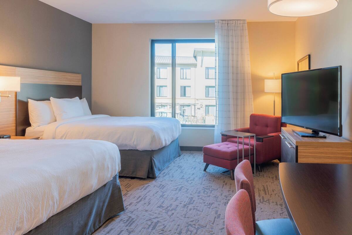 A hotel room with two beds, a TV, a desk, and a window with a view of buildings.