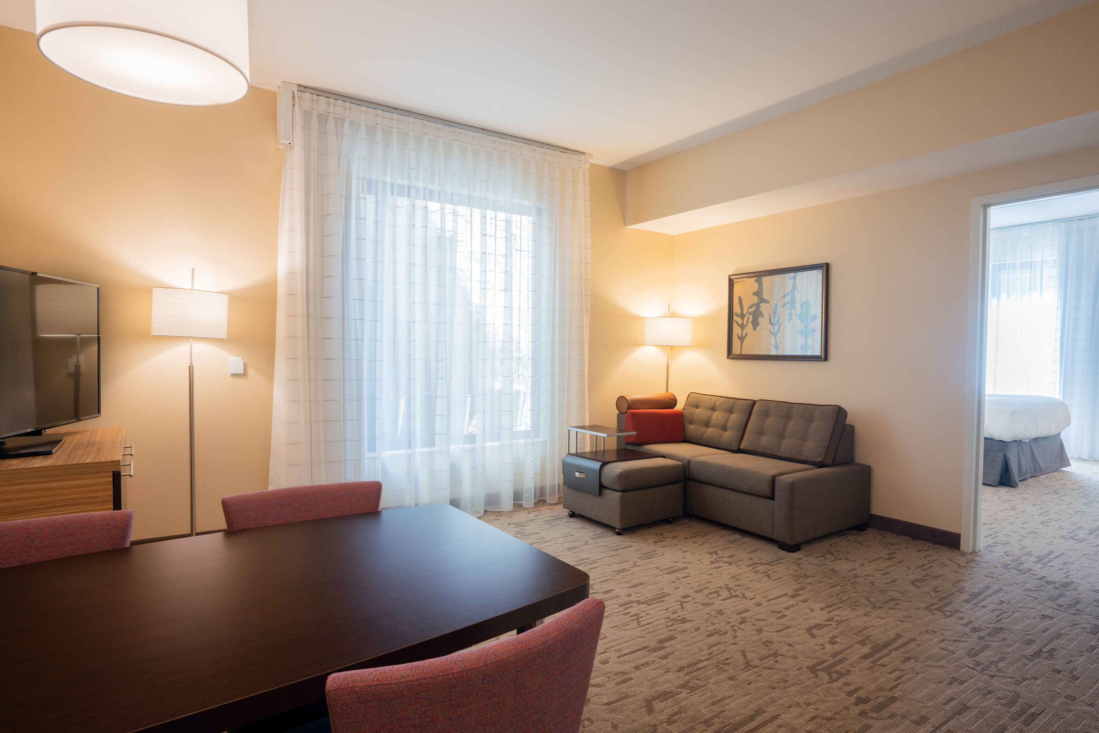 A hotel room with a dining table, chairs, a sofa, a TV, lamps, and a painting on the wall.