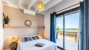 A bedroom with a bed, bedside lamps, a wall-mounted air conditioner, a sunburst mirror, and a view of the beach through a sliding glass door. - Villa Oceana exceptional property sea view from every room (Ondres)