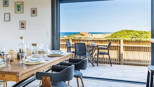 A dining area with a view of the beach, featuring a wooden table set with plates, glasses, and a bottle. - Villa Oceana exceptional property sea view from every room (Ondres)