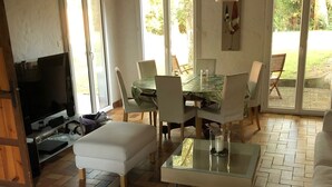 A cozy living room with a white sofa, a glass coffee table, and a TV. There is a dining area with a table and chairs, and large windows offering a view of the outdoors. - Beautiful style house near the golf course (Soorts-Hossegor)
