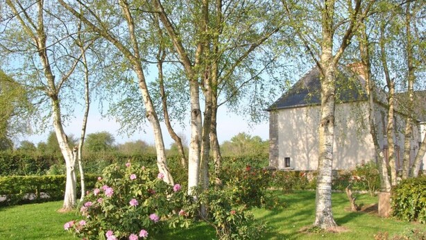 Een stenen gebouw met een schuin dak, omgeven door een verzorgde tuin met bloeiende struiken en bomen.