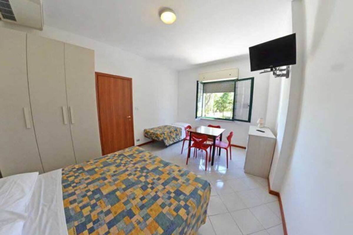 A bedroom with a bed, a desk, a chair, a TV, and a window.