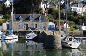 A harbor with boats docked, a stone building with blue doors, and houses in the background.