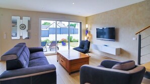 Een moderne woonkamer met een bank, fauteuil en salontafel, een aan de muur bevestigde televisie, en een glazen schuifdeur naar een terras.