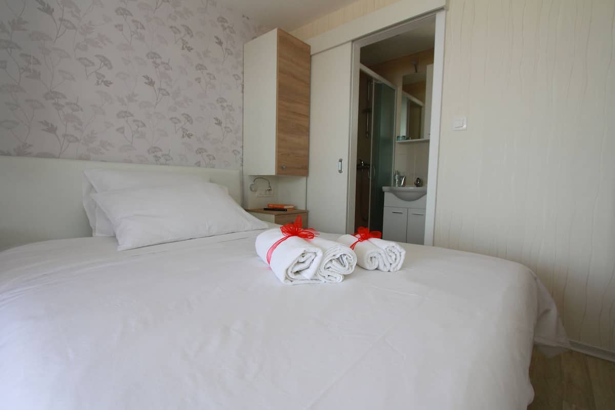 Una cama bien tendida con sábanas blancas y detalles en rojo, un armario de madera, un baño con lavamanos y espejo, y un estampado de papel tapiz floral.