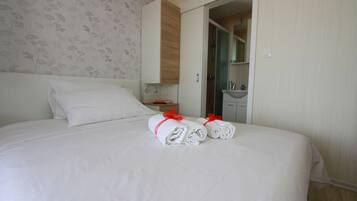 Una cama bien tendida con sábanas blancas y detalles en rojo, un armario de madera, un baño con lavamanos y espejo, y un estampado de papel tapiz floral.
