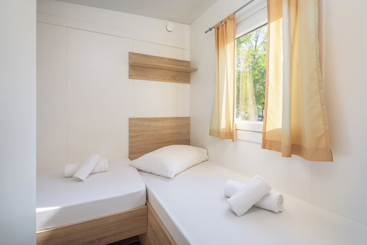 Una habitación pequeña y limpia con dos camas, cabeceros de madera y una ventana con cortinas translúcidas.