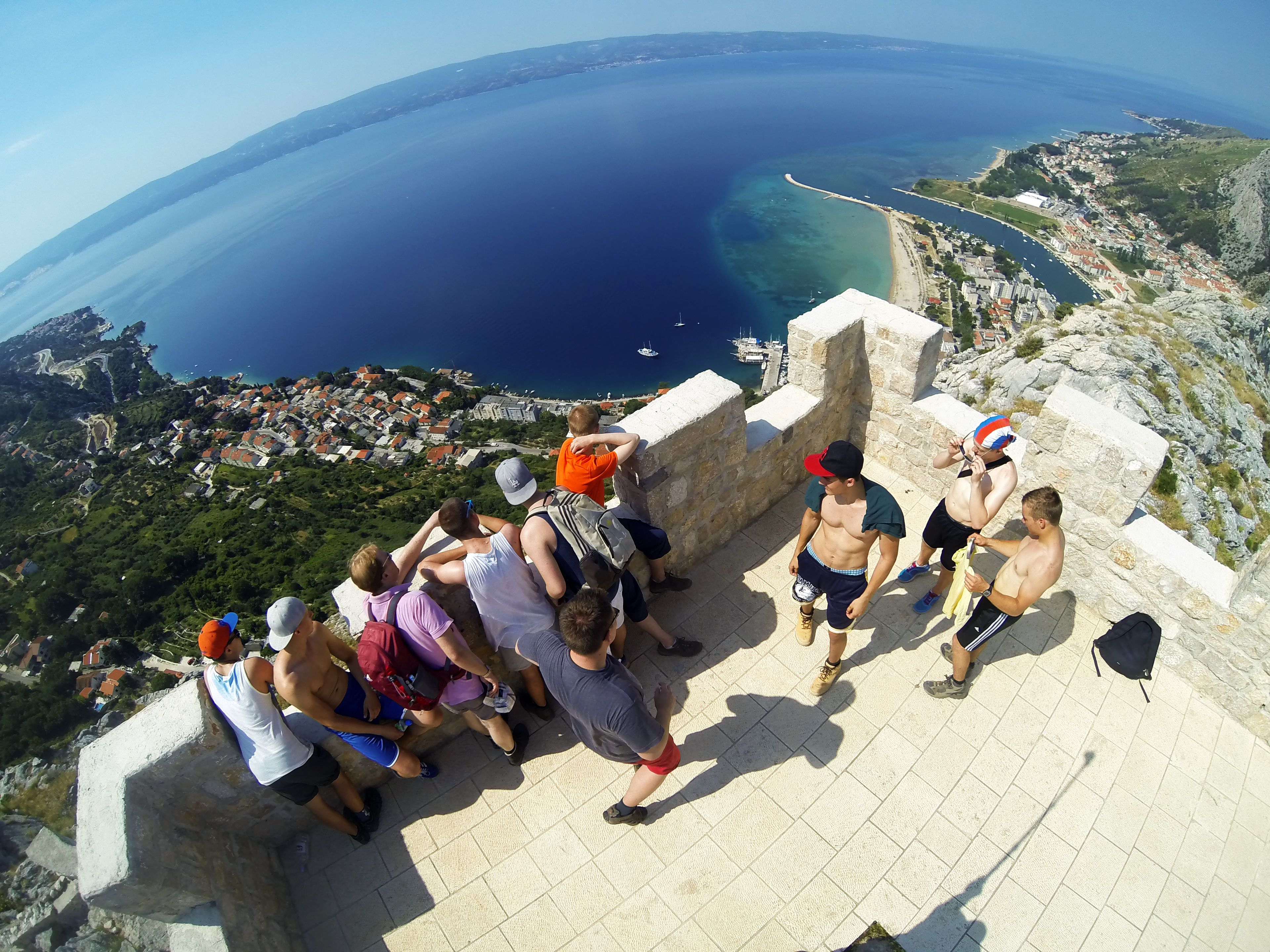 Un groupe de personnes se tient sur une plateforme en pierre, surplombant une ville côtière, d’où l’on a une vue dégagée sur la mer et les bateaux.