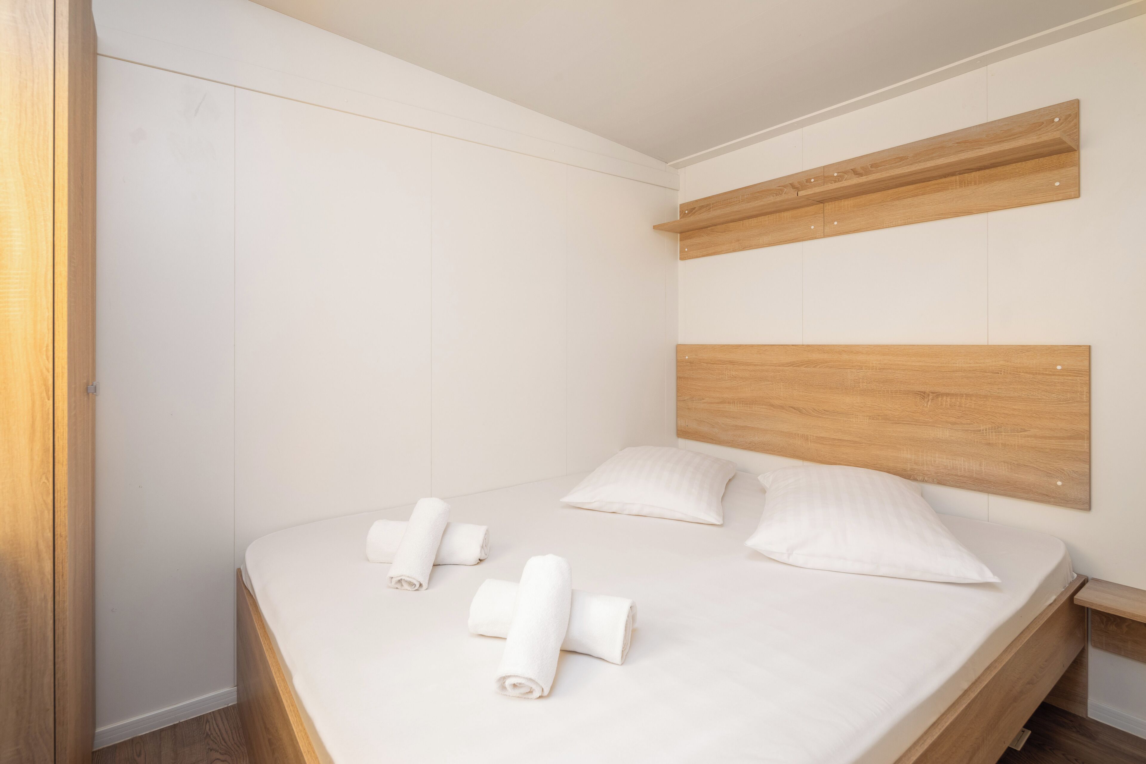 Ein ordentlich bezogenes Bett mit weißen Leinen und Kissen, ein Holz-Kopfbrett und ein Nachttisch aus Holz mit einer Lampe.