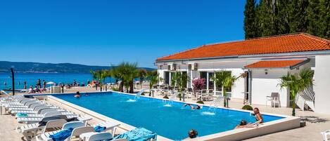 A swimming pool with lounge chairs, a building with a red roof, and a view of the sea and hills.