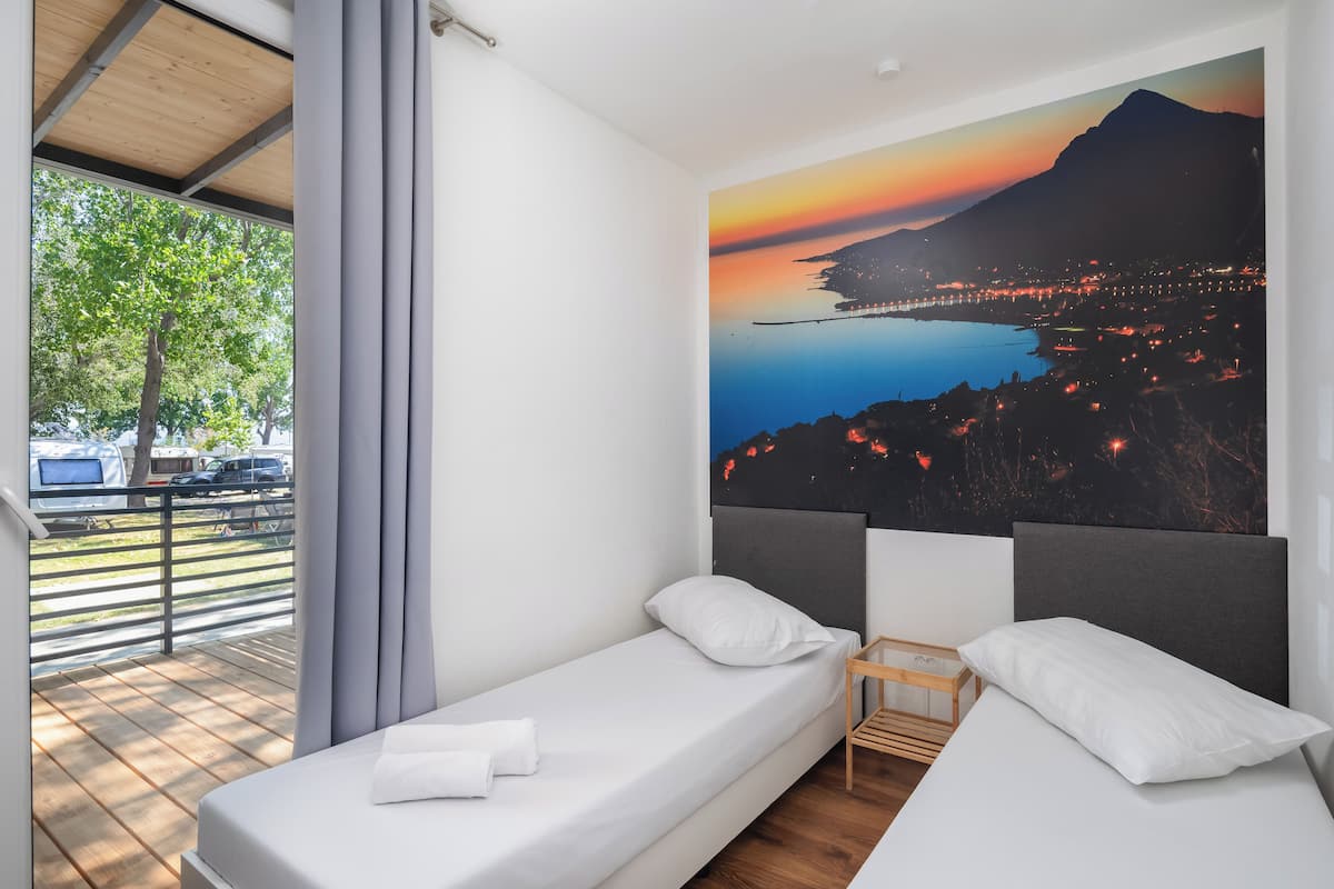 Una cama individual con cabecera de madera, una mesita de noche pequeña y vista a un paisaje urbano al atardecer.