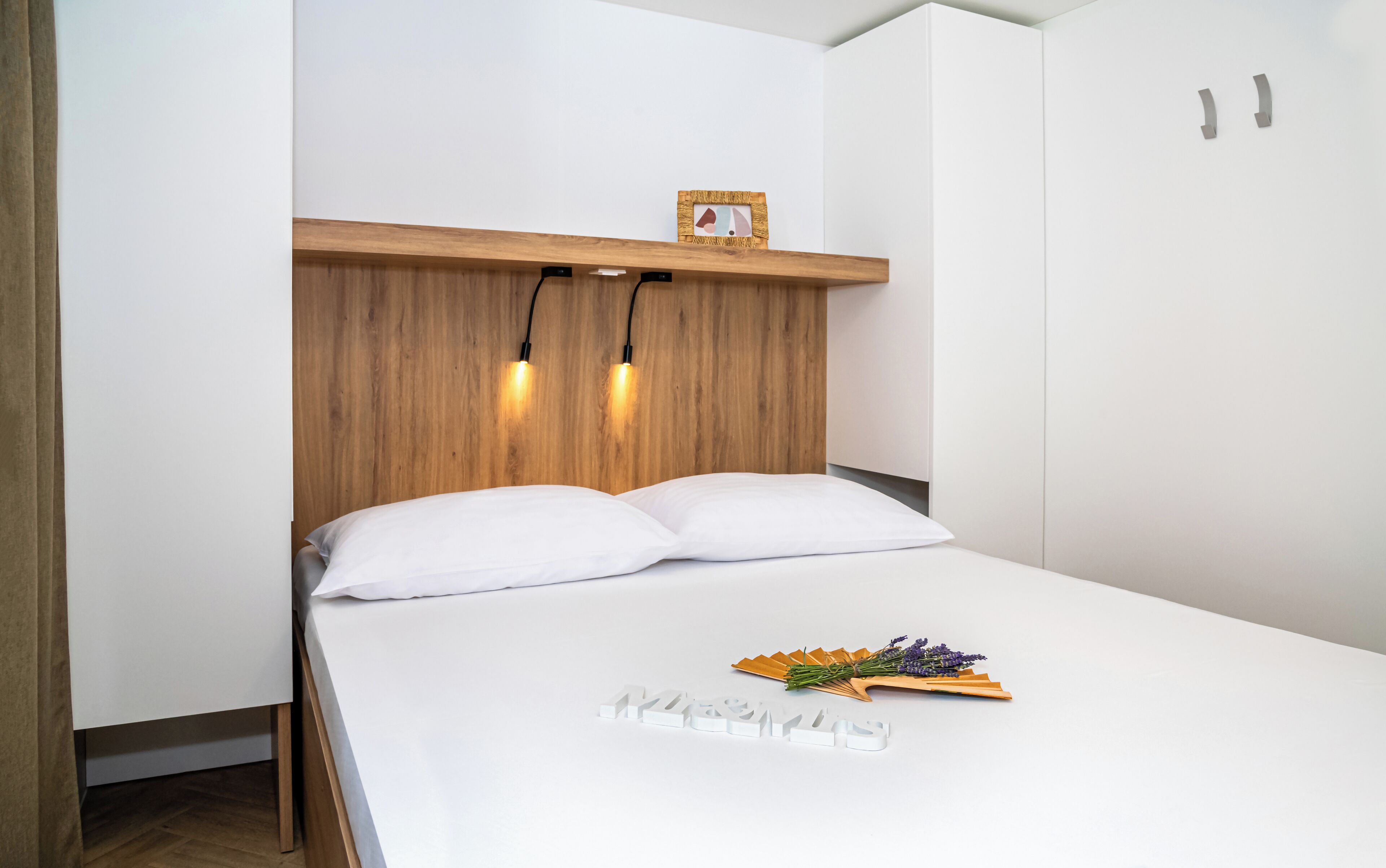 A neatly made bed with white linens and pillows, a wooden headboard with two wall-mounted lights, and a small framed picture above the headboard.