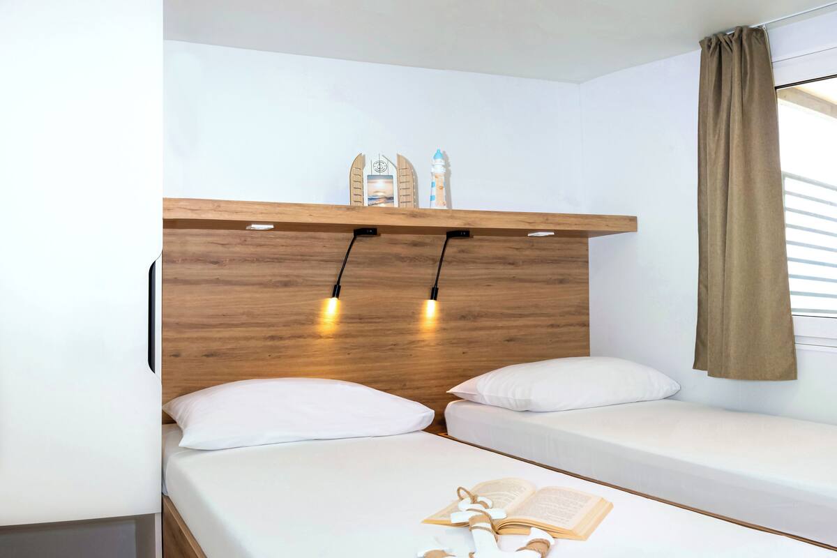 Un dormitorio con dos camas individuales, cabecera de madera, lámparas de noche, estante con objetos decorativos y una ventana con cortinas.
