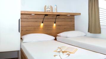 A bedroom with two single beds, a wooden headboard, bedside lamps, a shelf with decorative items, and a window with curtains.