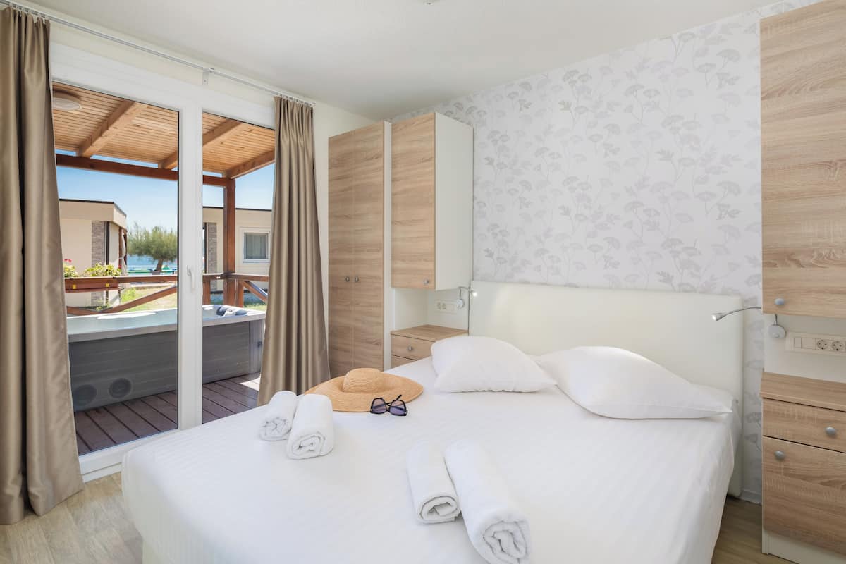 Habitación de hotel con cama, cabecero de madera, ventana grande con cortinas y mesita de noche de madera, con vistas a una zona de piscina exterior.