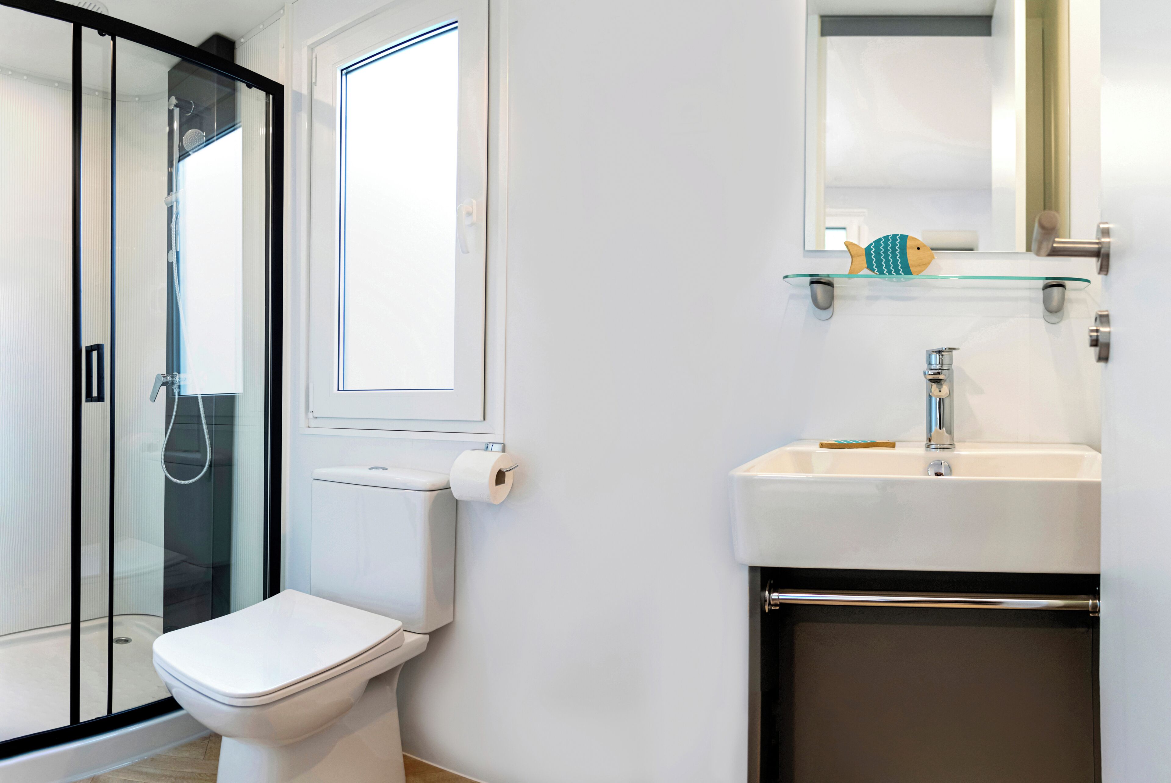 A modern bathroom with a glass shower enclosure, a toilet, a sink with a mirror above, and a small shelf with a toothbrush.
