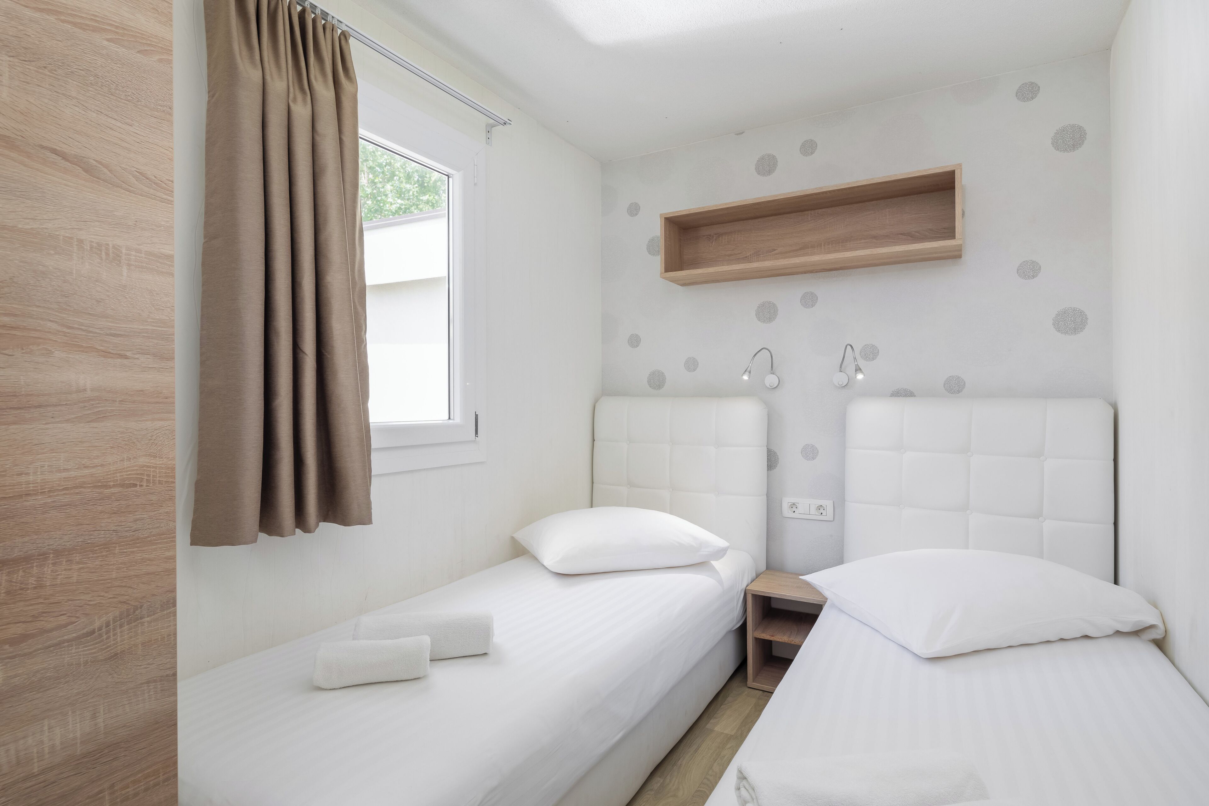 Habitació petita i moderna amb dos llits individuals, una prateleira de fusta i una finestra amb cortines.