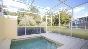 Una piscina techada con techo de vidrio, rodeada por un muro blanco y con vistas a árboles y un edificio al fondo.